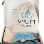 A box with the aerial hammock and rigging equipment. The box includes a beige bag with “Uplift Active” on it and a tie dye hammock. 